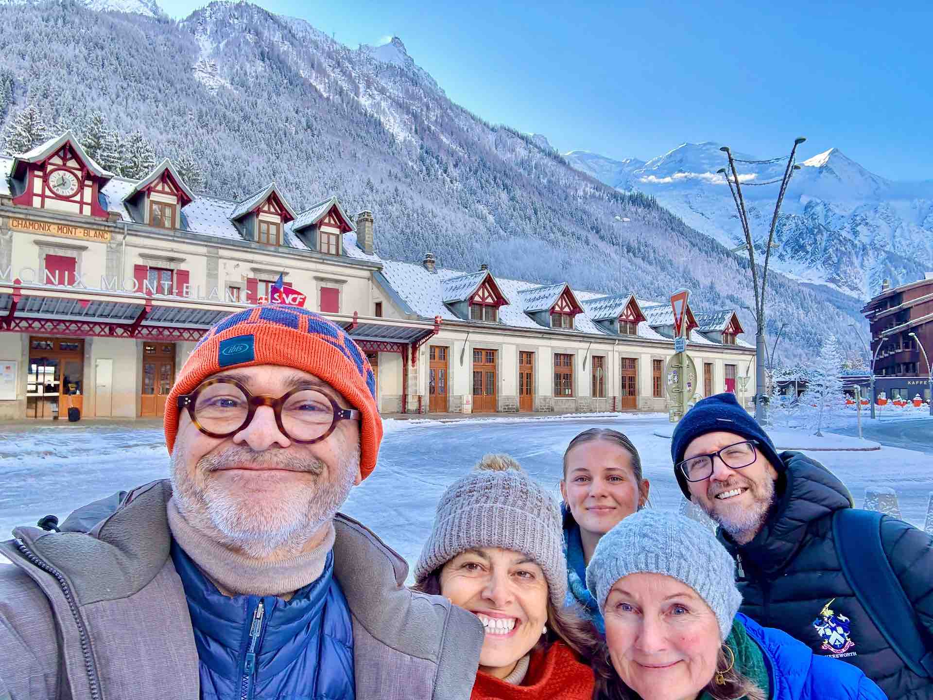 Chicca with husband Arnaud and friends in front of the Chamonix-Mont-Blanc train station, with snow-capped Alpine peaks in the background.