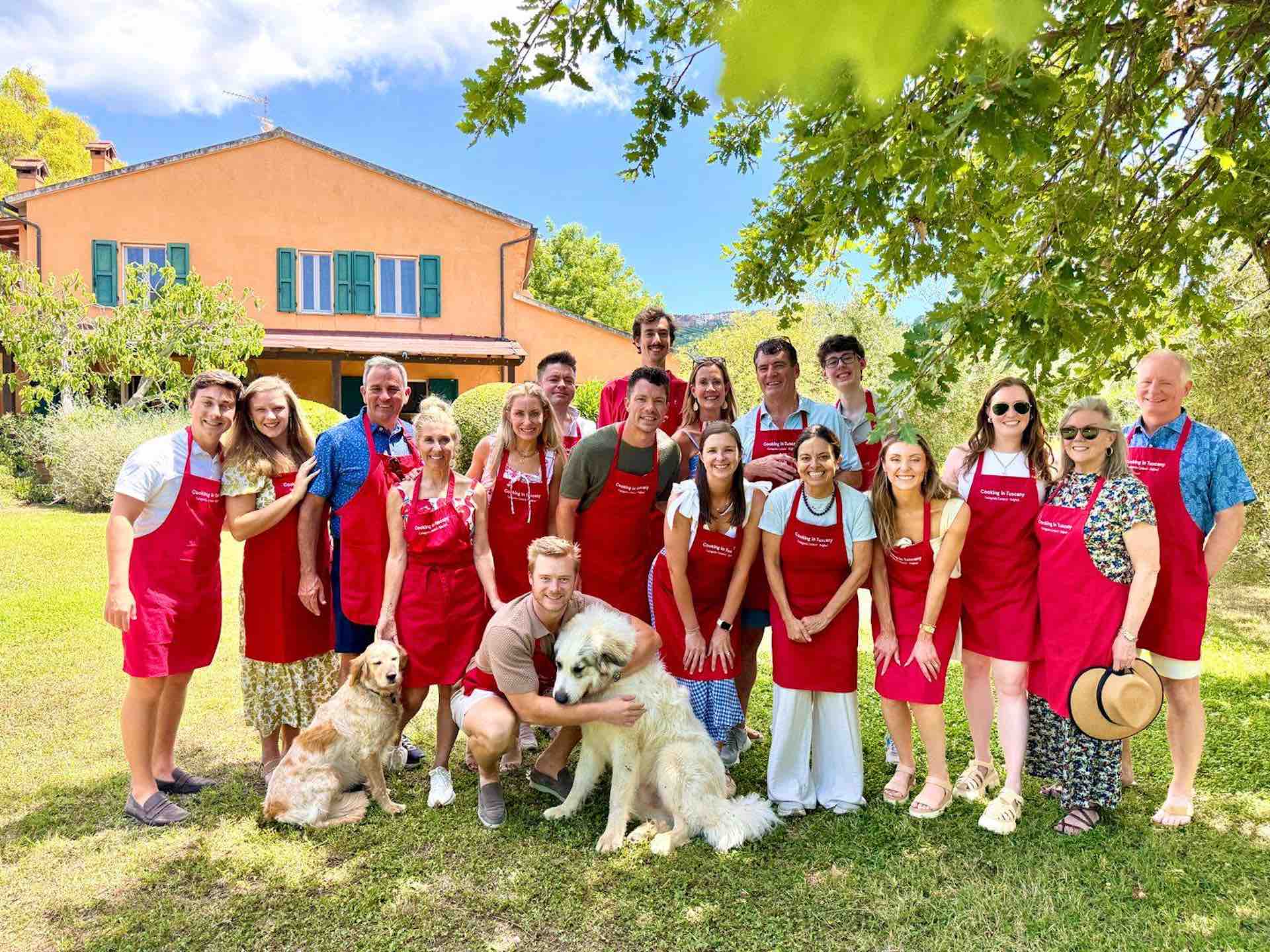 Group of wine tour guests in red aprons posing with two golden retrievers in front of a Tuscan farmhouse