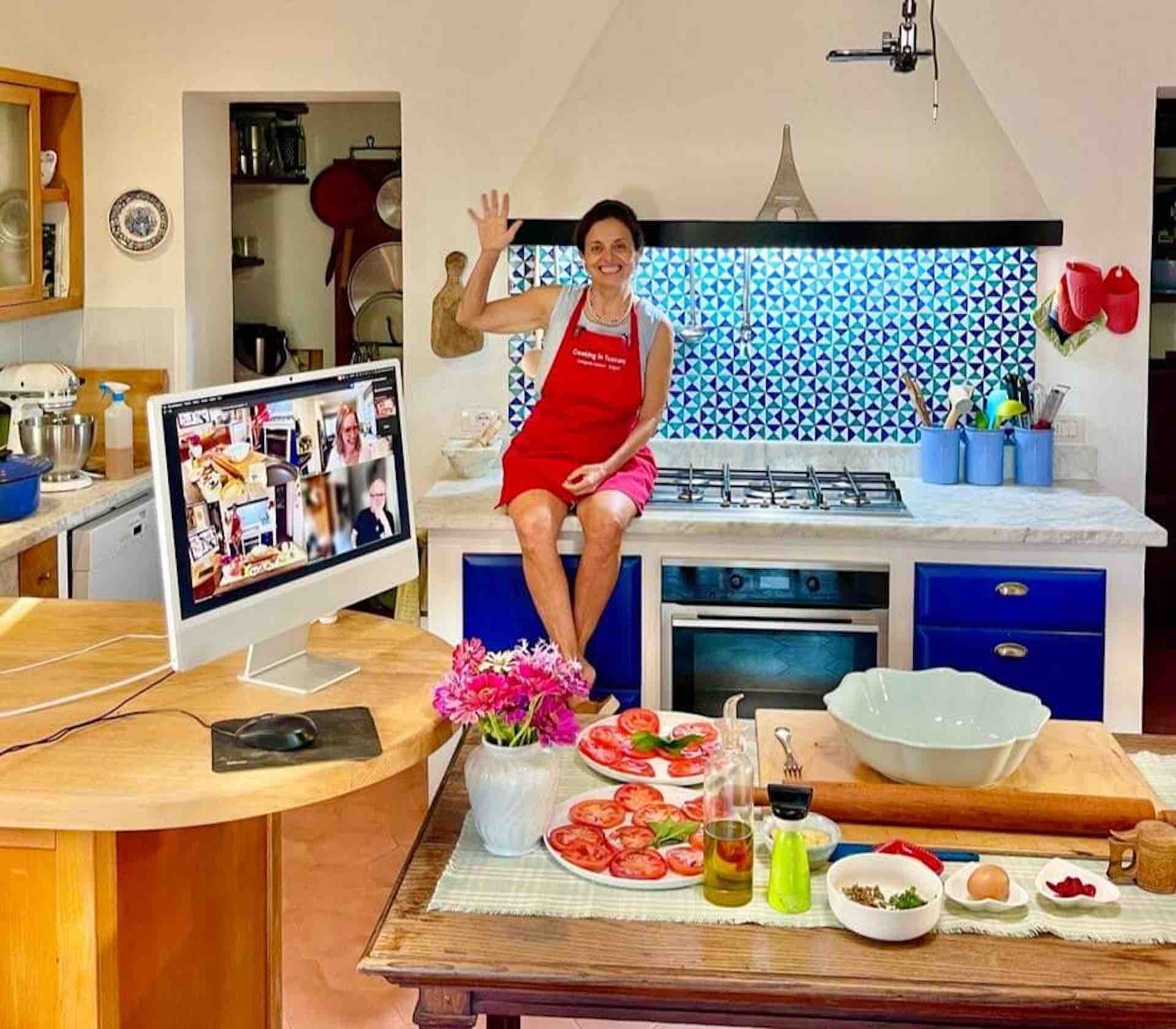 Chicca Teaching a Live Online Cooking Class Chicca waving from her Tuscan kitchen counter beside a laptop showing class participants, with fresh tomatoes on the table