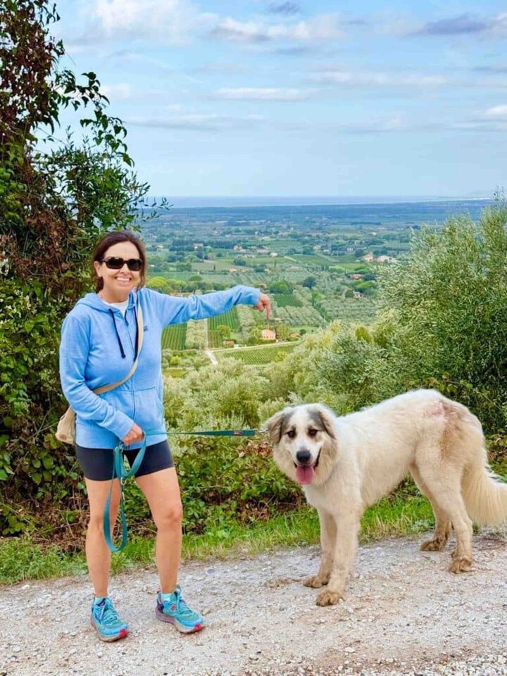 Chicca and Her Dog Overlooking the Tuscan Valley Chicca in a blue jacket and sunglasses standing on a hillside path with a large white dog, pointing toward the green Tuscan valley below