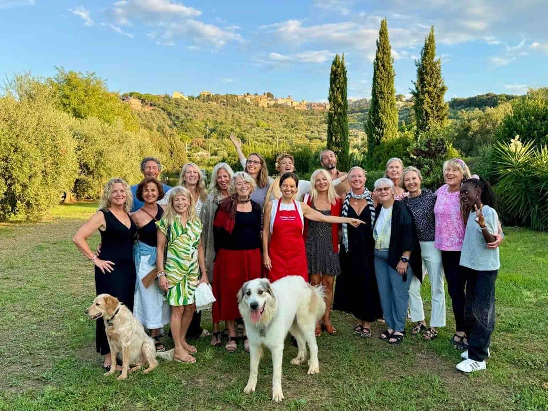 Cooking Class Group in the Tuscan Garden Group of smiling cooking class guests standing together in a Tuscan garden at sunset, with Chicca in a red apron and two dogs in front