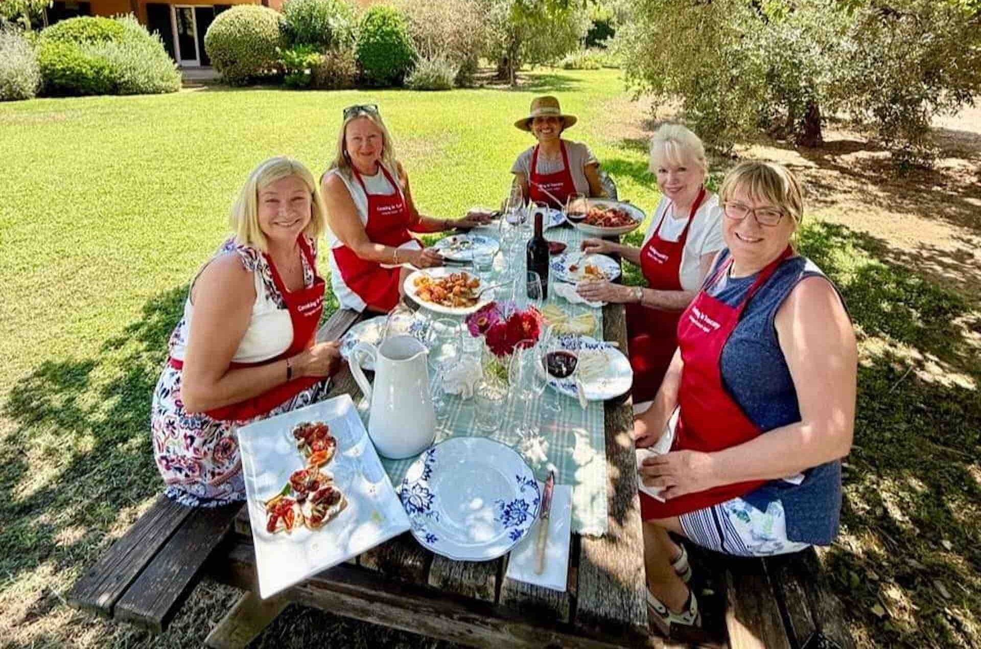 Cooking Club Season 21 Group Celebration Five women in red Cooking in Tuscany aprons seated at an outdoor table with plates of food, celebrating Season 21